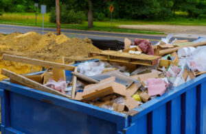 A blue dumpster filled with construction debris, cardboard, wood, and various discarded materials sits near a road in CA, ideal for Junk Removal San Francisco County projects amid piles of dirt and trees in the background.