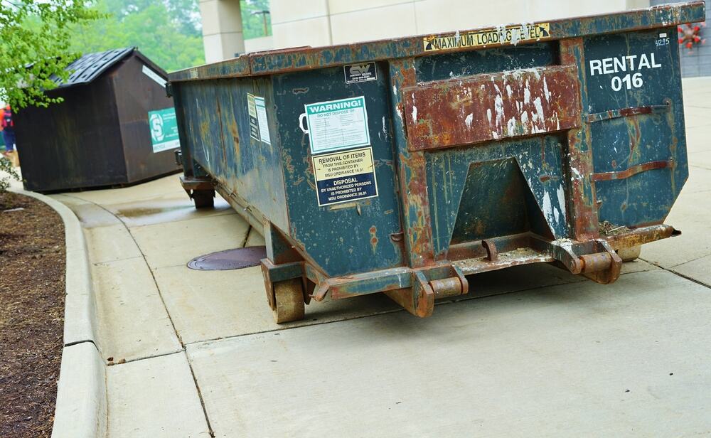 A large, rusty metal dumpster labeled "RENTAL 016" sits on a concrete driveway near a building in CA, with another smaller dumpster in the background and some greenery on the left, perfect for Junk Removal San Francisco County.