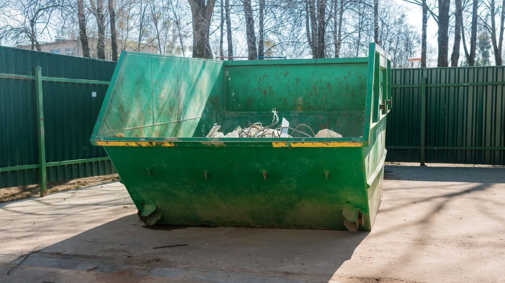 A large green dumpster sits on a concrete surface, holding assorted junk and broken wires, akin to the typical sights in San Francisco County. Encircled by green metal fences and leafless trees under a clear sky, it seems ready for efficient junk removal.