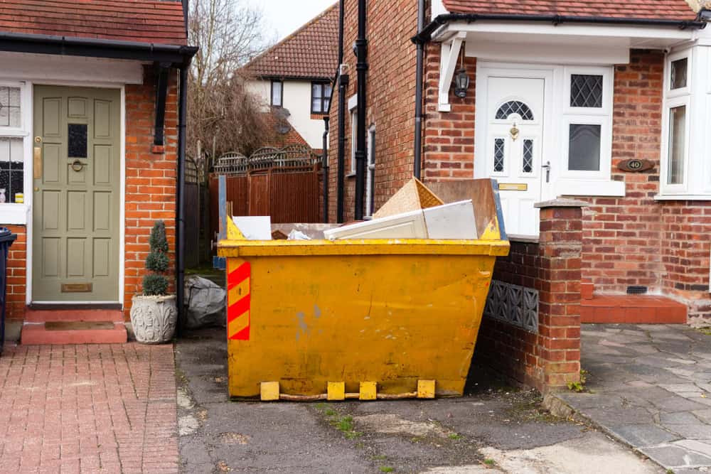 A yellow dumpster filled with construction debris sits in the driveway between two brick houses with red-tiled roofs. One house boasts a green door, the other a white one. This suggests renovation work, perhaps aided by a convenient dumpster rental in San Francisco County, CA.