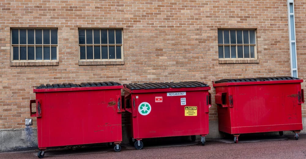 Three red dumpsters, perhaps from a junk removal service in San Francisco County, are lined up against a brick building with small, barred windows. One has a recycling symbol. The ground is paved with a subtle texture, and there's a white pipe on the right side of the wall.