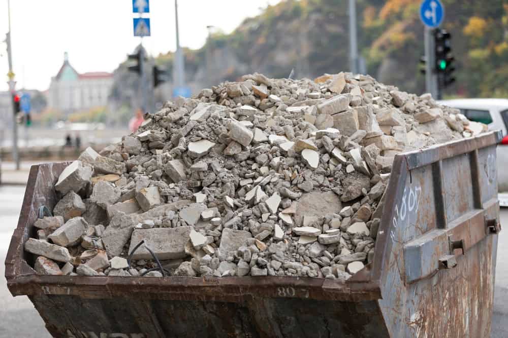 A large metal dumpster, emblematic of junk removal in San Francisco County, is filled with broken concrete rubble on a street. Traffic lights and cars form the backdrop, framed by trees and a distant building under the cloudy CA sky.