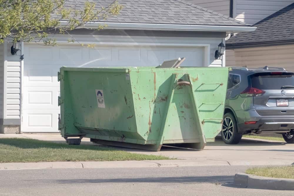 A large green dumpster rental from San Francisco County is placed on the driveway in front of a white garage door. A grey SUV is parked nearby. The suburban house has light-colored siding and asphalt shingles. A small tree is visible on the left.