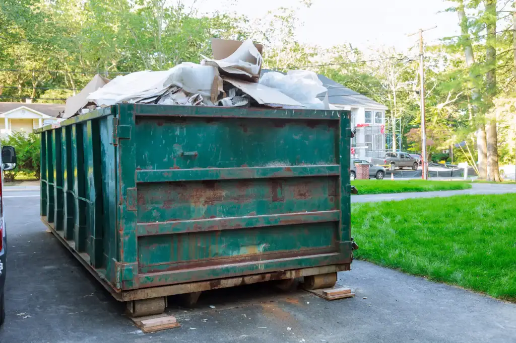 A large green dumpster filled with cardboard, plastic, and other debris sits on a driveway in a CA residential neighborhood, highlighting the need for Junk Removal San Francisco County services among trees and houses in the background.