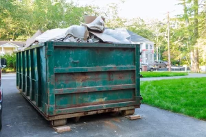 A large green dumpster filled with cardboard, plastic, and other debris sits on a driveway in a CA residential neighborhood, highlighting the need for Junk Removal San Francisco County services among trees and houses in the background.