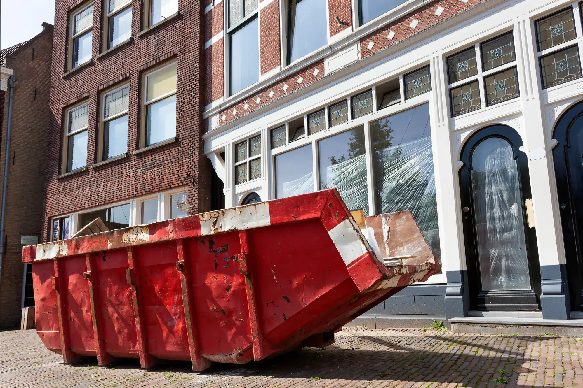 A large red dumpster sits on a cobblestone street in front of brick buildings, with one building's windows covered in plastic sheeting&mdash;classic signs of renovation or Junk Removal San Francisco County, CA.